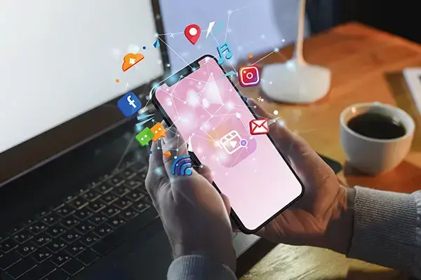 Hands holding a smartphone with vibrant social media icons, connecting to a laptop in a modern work setting.