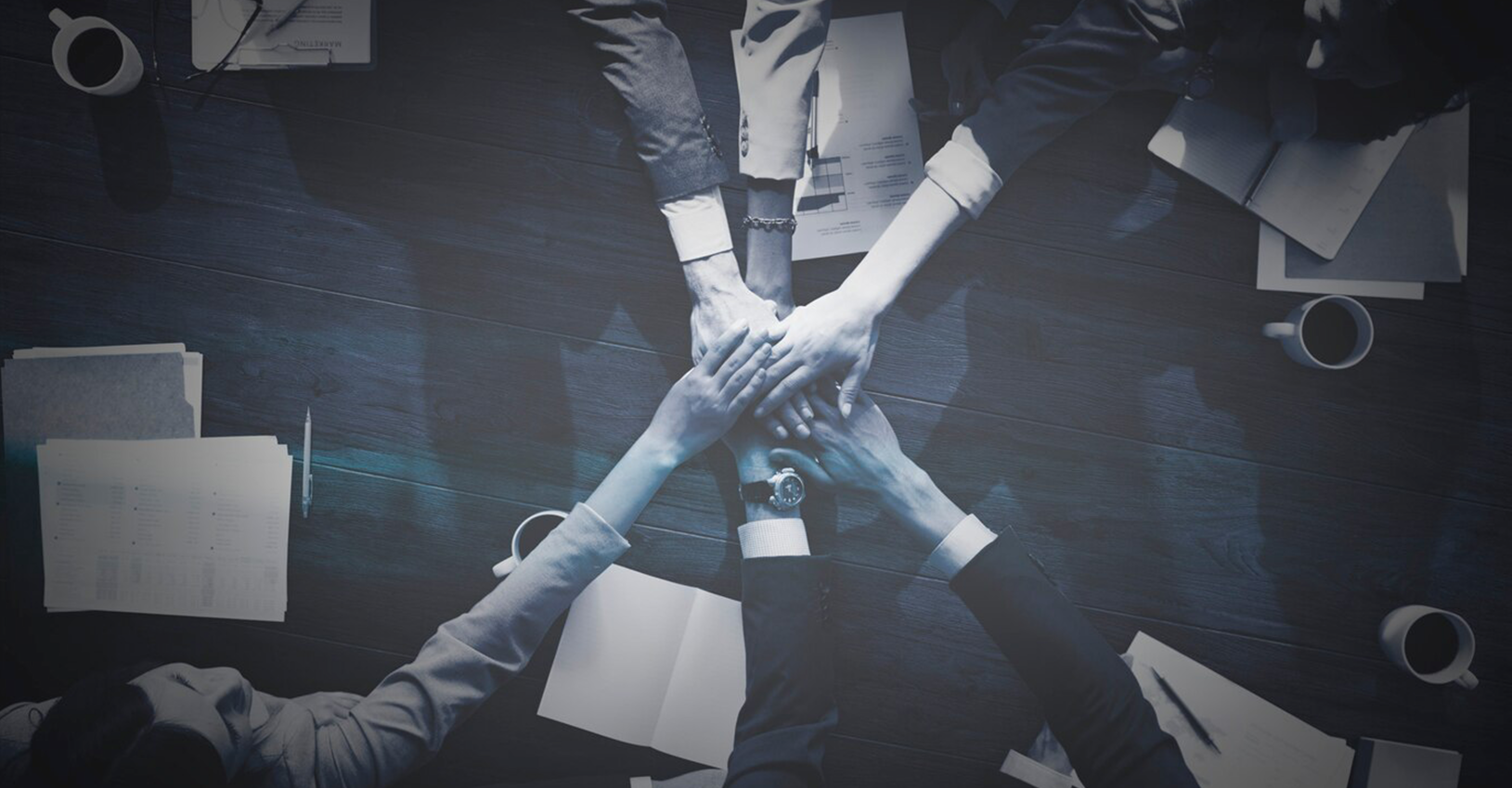 Top view of business team placing hands together in a show of unity and teamwork on a dark wooden table, symbolizing collaboration and partnership.